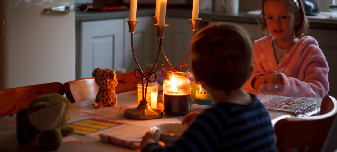 The power has gone out and two children are sitting together at a table at home with candles in front of them for light